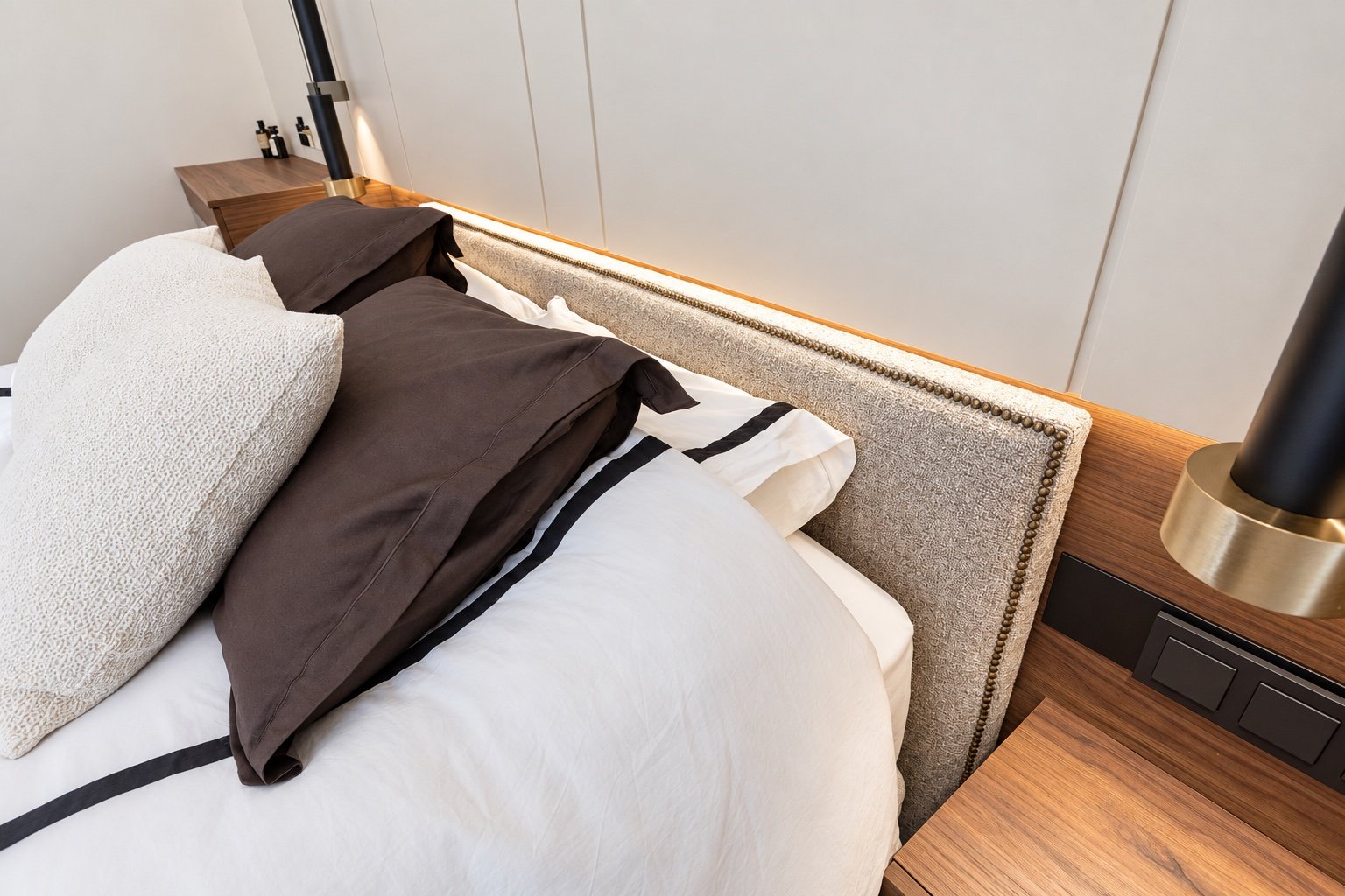 Detalle de cabecero tapizado, ropa de cama blanca y marrón y mesita de madera