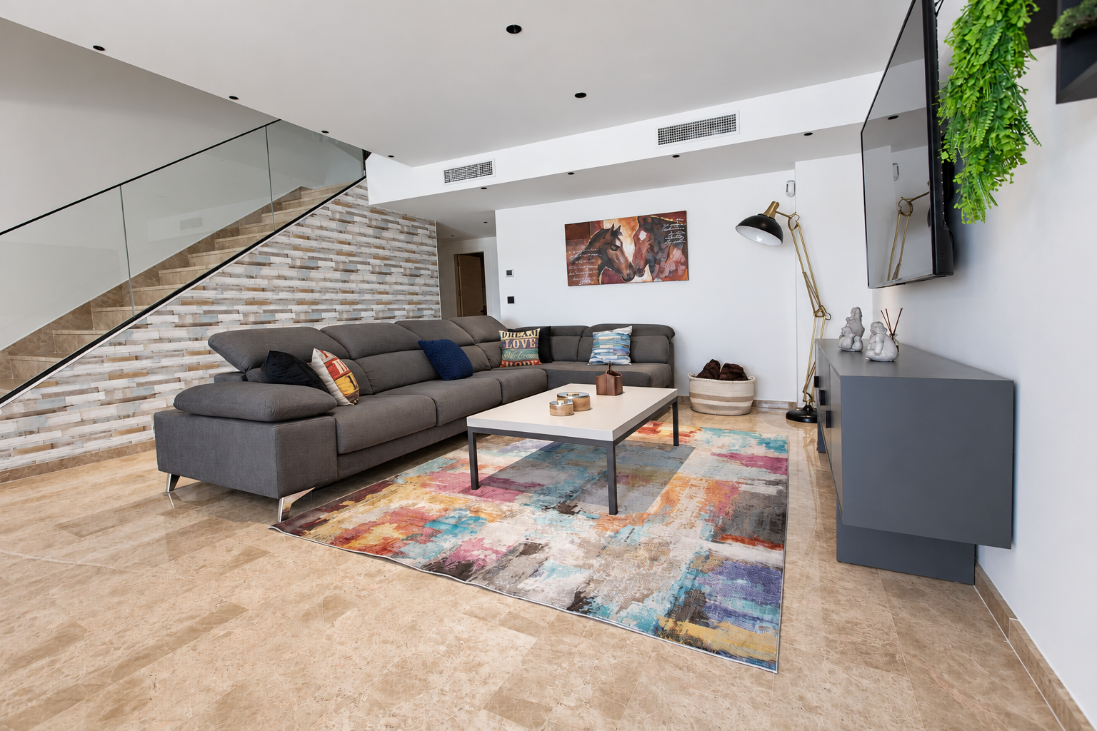 Salón con sofá gris, alfombra de colores y escalera con barandilla de cristal
