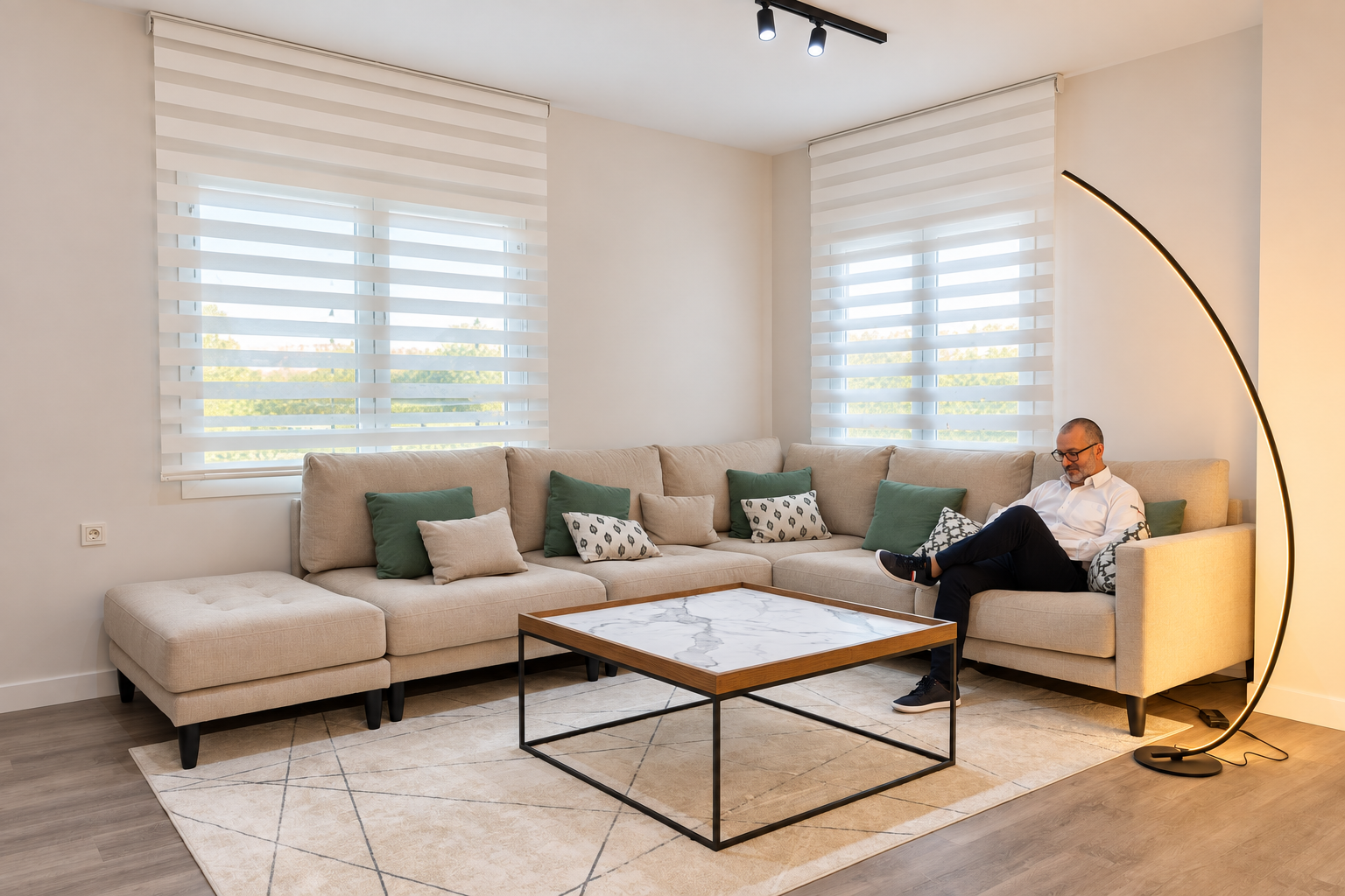 Salón con sofá beige, cojines verdes y una persona sentada junto a una lámpara de arco
